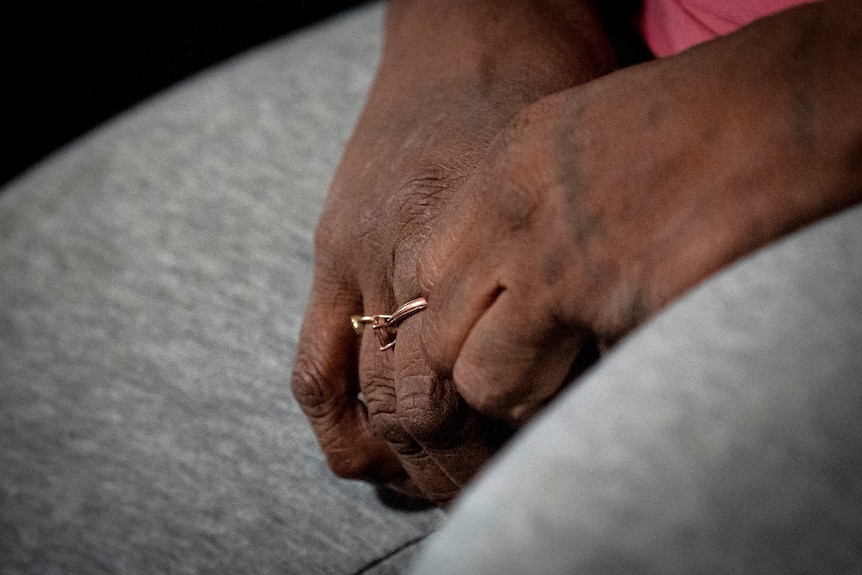 A close-up of an older Indigenous woman's hands resting on grey pants, with a ring on one of her fingers.