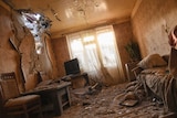The interior of an apartment where light streams through a hole in the wall, with broken glass and rubble on the floor.