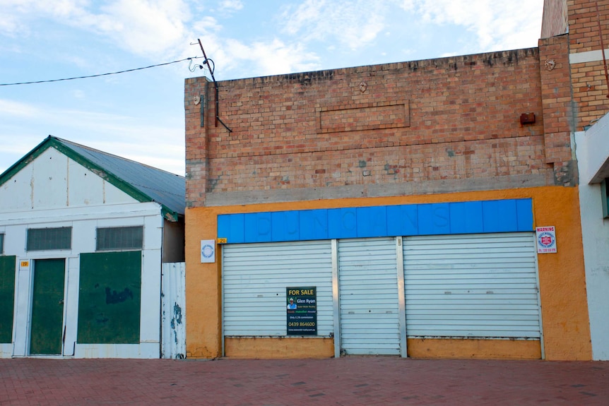 Building shuttered up with a for sale sign on the front, another boarded up building beside it