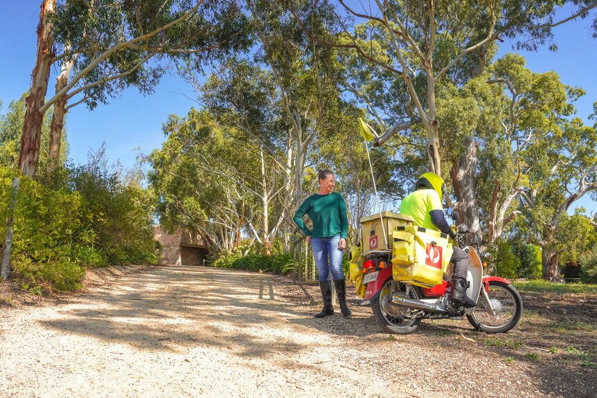A woman stands at the end of a long driveway chatting with an Australia Post employee delivering mail on a postie bike.