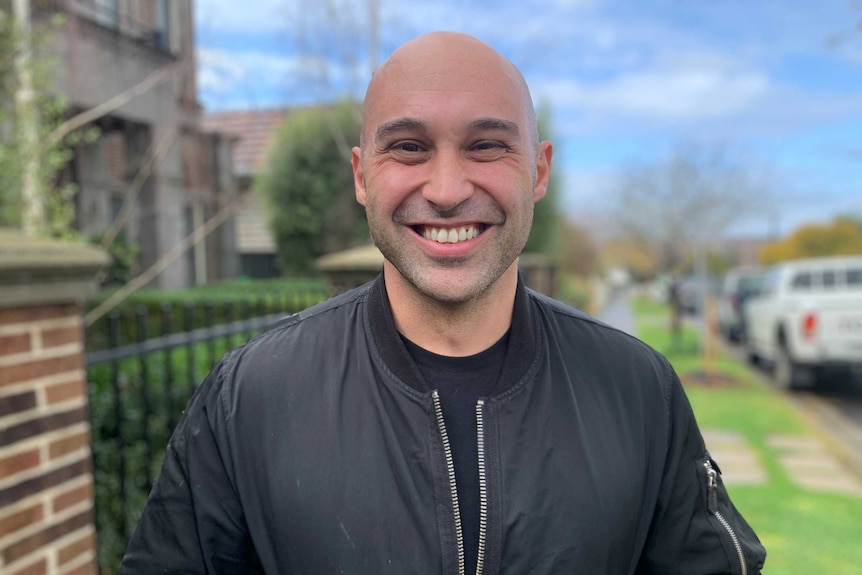 Chef Shane Delia, wearing a black shirt and bomber jacket, smiles broadly at the camera.