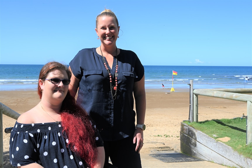 A woman sits in a wheelchair next to a lady with blonde hair smiling at Neilsen's Beach in Bargara.