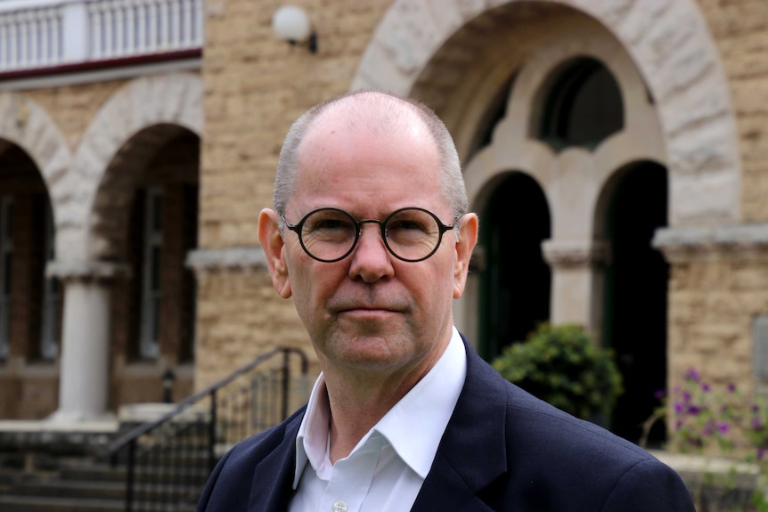 A balding man wearing circular glasses stands in front of a heritage-style sandstone building.