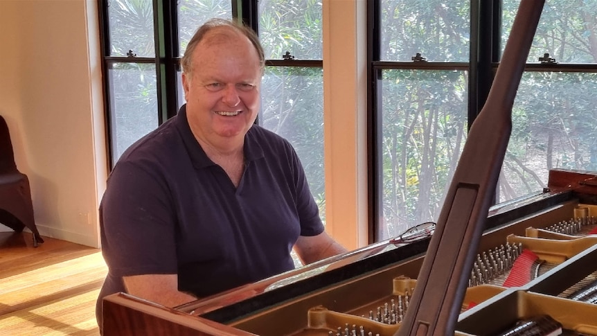 A balding older man in dark polo shirt, sits in a sunlit room at a piano and smiles.
