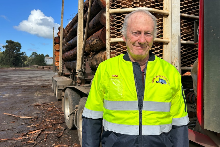 An older man in a high-vis shirt stands next to a truck loaded with logs.