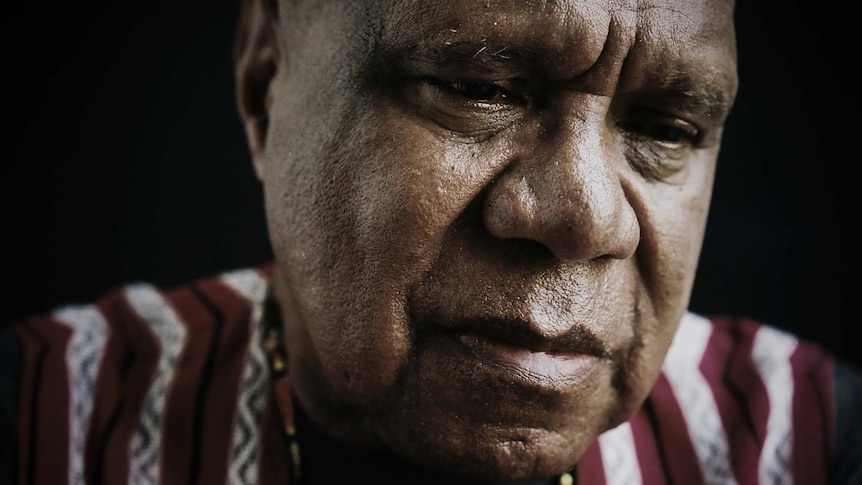 Archie Roach is wearing a striped shirt and sitting under dim lights. He is looking down and his expression is serious.