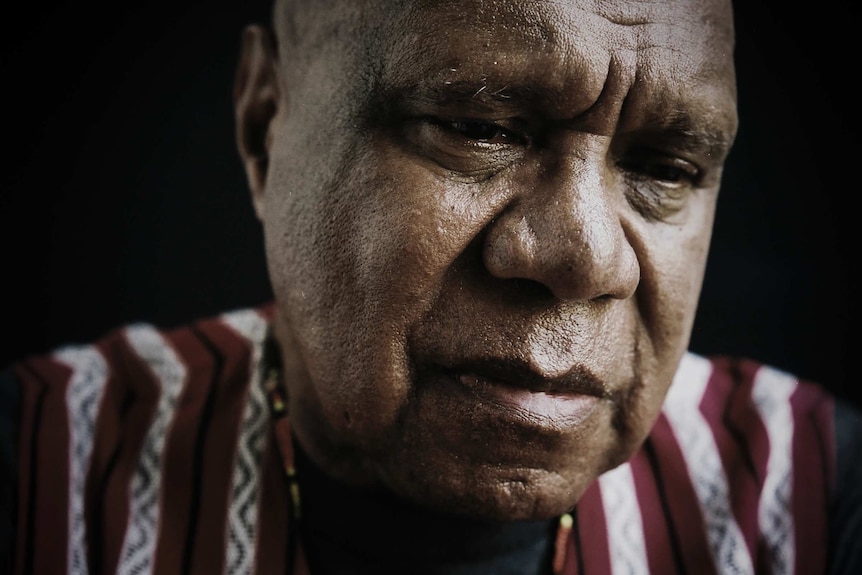 Archie Roach is wearing a striped shirt and sitting under dim lights. He is looking down and his expression is serious.