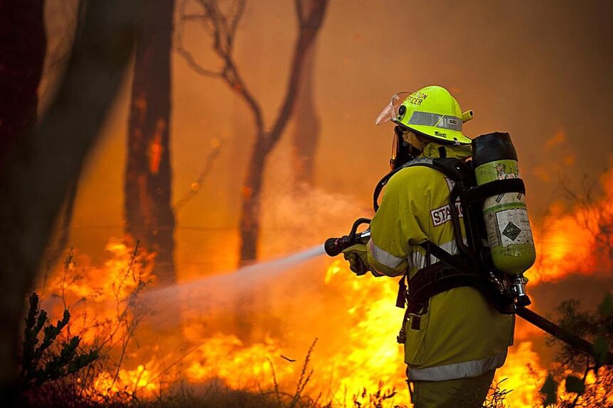 A firefighter wearing breathing apparatus battles a blaze with flames in the background.