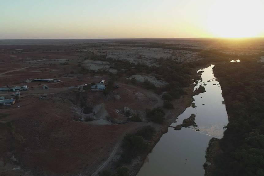An aerial view of buildings in the outback overlooking a river, against a backdrop of trees and plains, and a setting sun.