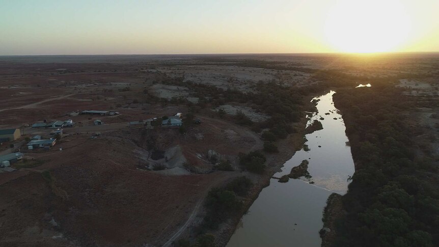 An aerial view of buildings in the outback overlooking a river, against a backdrop of trees and plains, and a setting sun.