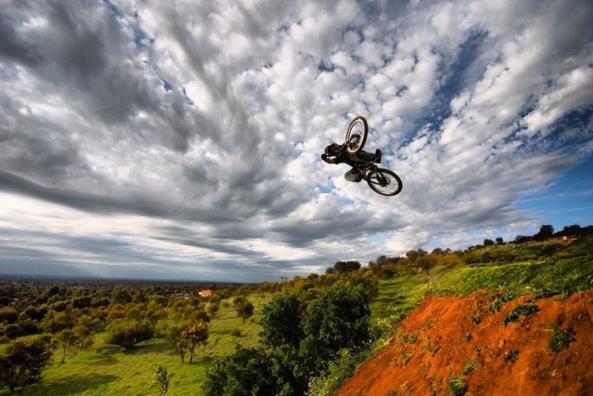 A man riding a bike high in the air over a jump with clouded sky and landscape as the backdrop