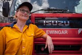 Woman in a rural fire brigade yellow uniform leans against a fire truck.