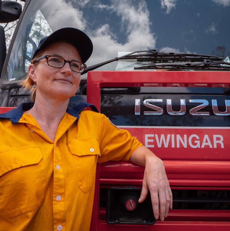 Woman in a rural fire brigade yellow uniform leans against a fire truck.