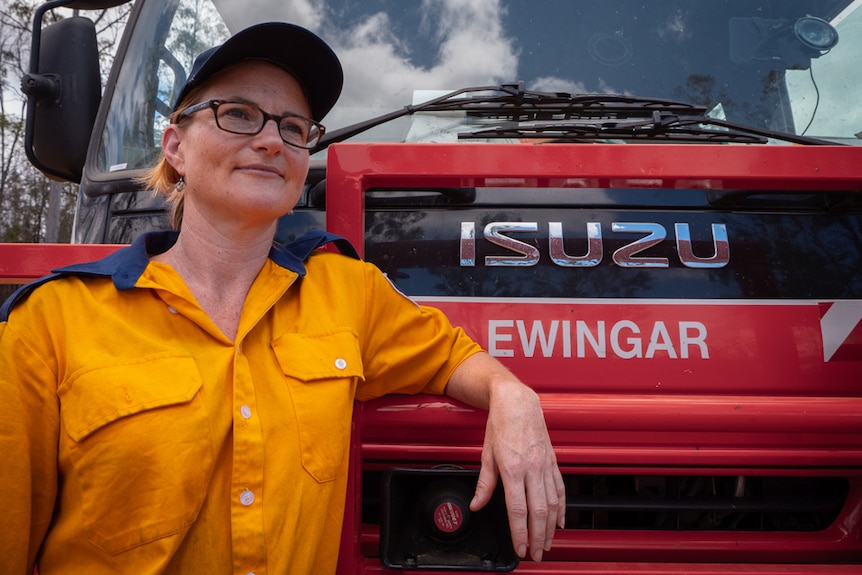 Woman in a rural fire brigade yellow uniform leans against a fire truck.