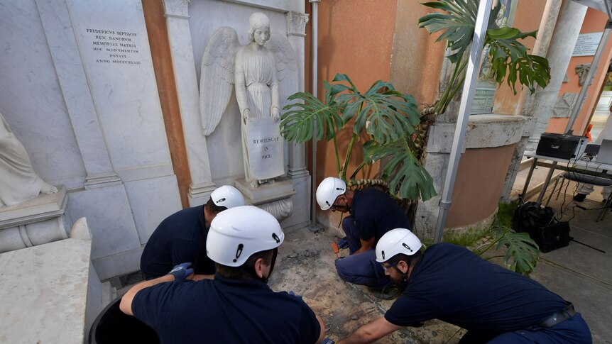 Four male workers in navy blue shirts and white helmets drill into a closed burial plot with a white angel towering over them.