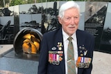 An elderly man with white hair stands in front of a war memorial while wearing a navy blazer adorned with military medals and ba