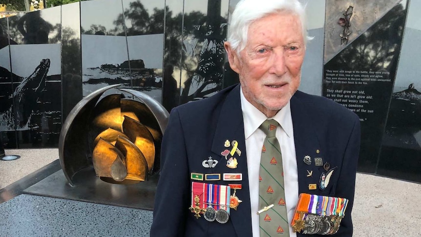 An elderly man with white hair stands in front of a war memorial while wearing a navy blazer adorned with military medals and ba