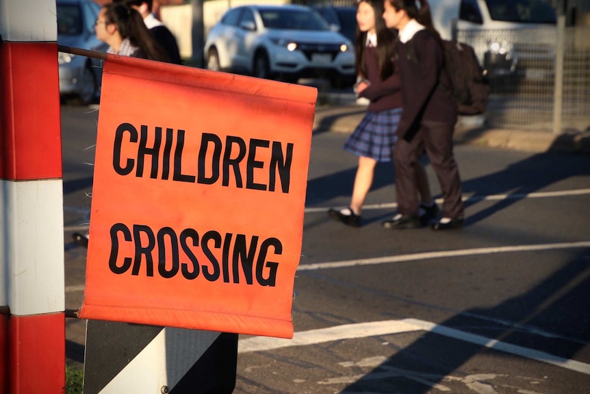 An orange children crossing sign with students walking across the road in the background.