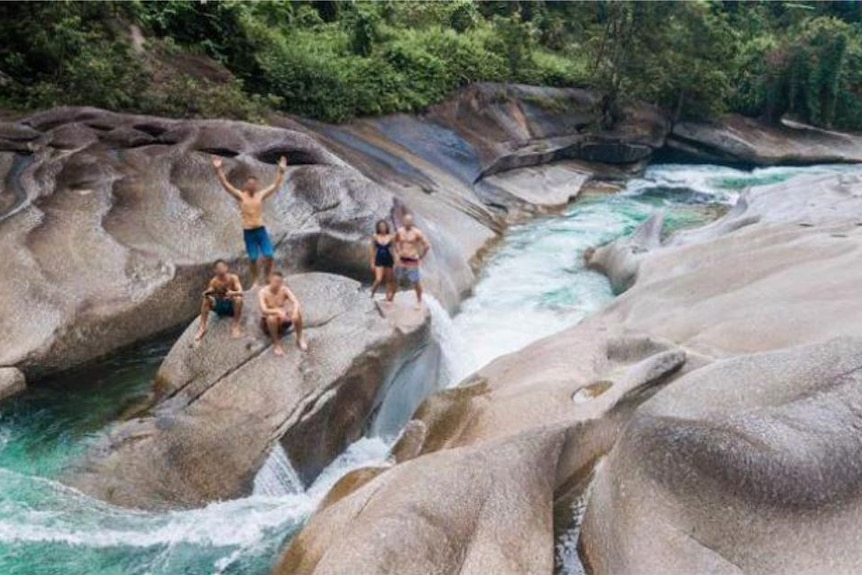 An aerial image of five people standing and crouching on a rock next to fast moving water.