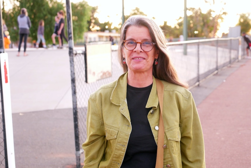 A woman in a green jacket and grey hair smiling in front of a skate park 