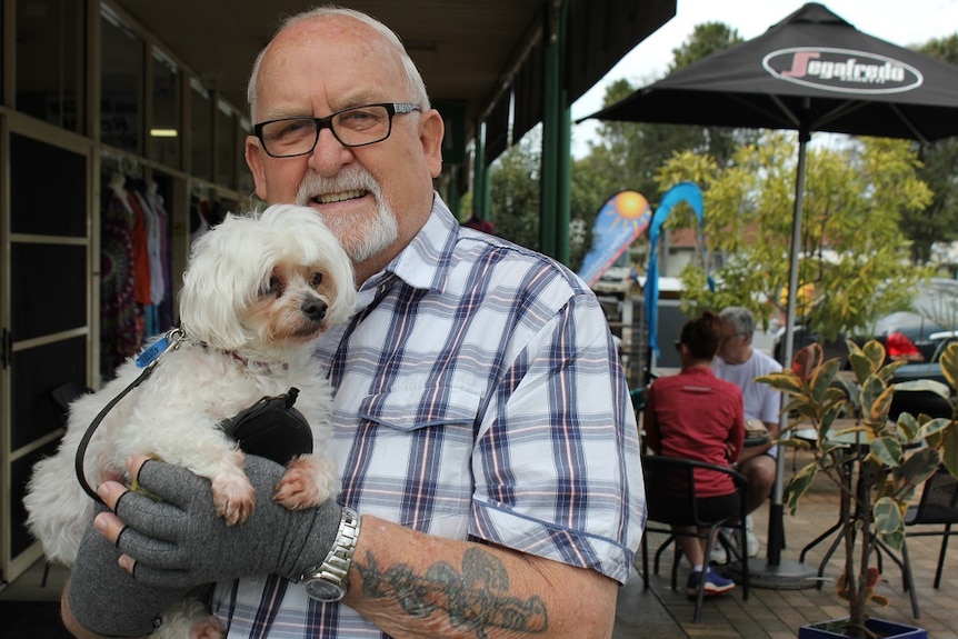 elderly man standing smiling with a white maltese dog on a street outside a cafe.