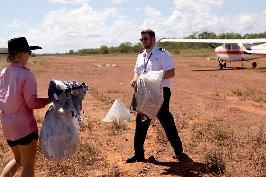 Woman walks towards pilot both carrying bags on a dirt airstrip. Plane in the background.