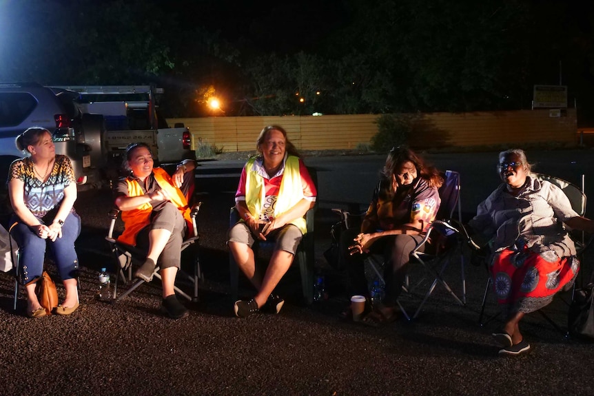 Woman sit in camping chairs at night time, with cars in the background.