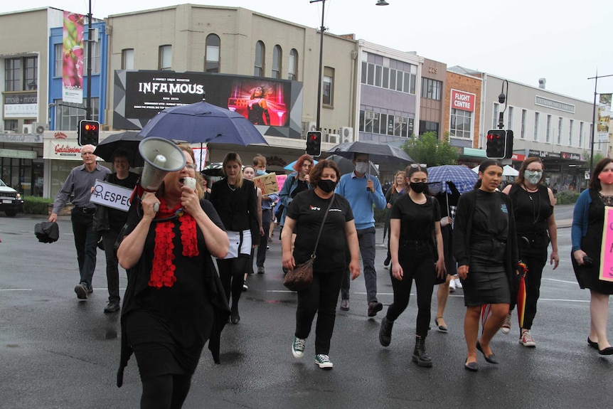 Women on megaphone crossing a road with dozens of women following holding signs and umbrellas.