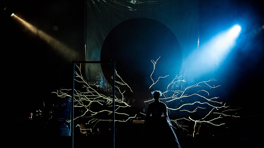 A silhouetted female figure on a set with dramatic spot lighting, a suspended frame and large tree branches with no leaves.