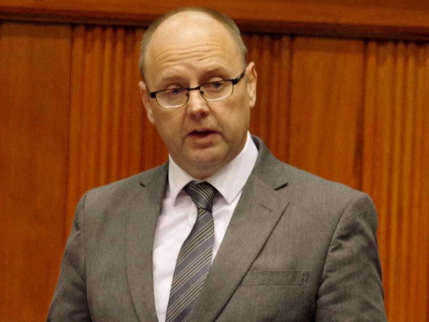 Barry Urban stands in Parliament, holding a piece of paper.