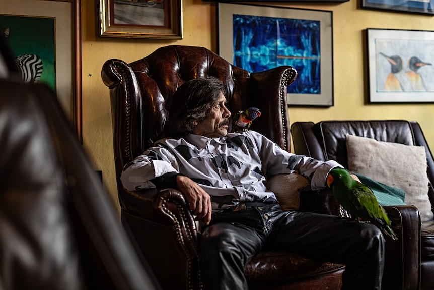 A man in white looks away from the camera, sitting in an armchair with a parrot perched nearby