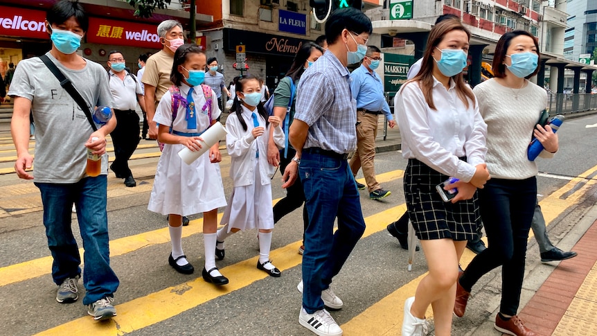 People wearing face masks cross a built up street at a yellow zebra crossing.