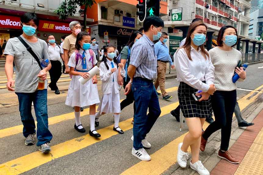 People wearing face masks cross a built up street at a yellow zebra crossing.