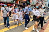 People wearing face masks cross a built up street at a yellow zebra crossing.