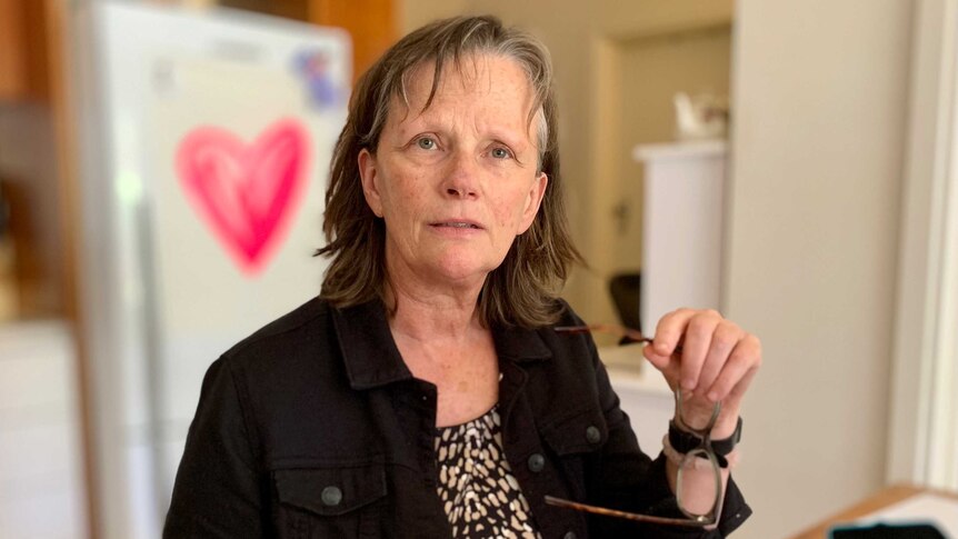 Dr Catherine Barrett sits at table holding her spectacles in a kitchen in a house.