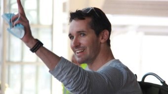 A man smiles and waves his left hand while holding a disposable face mask.