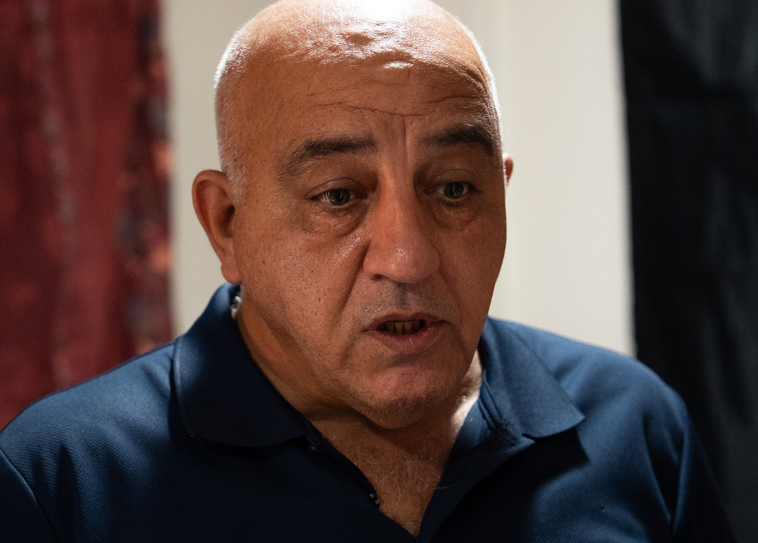 Un hombre calvo, de mediana edad y con camisa oscura, habla en una habitación de hotel.