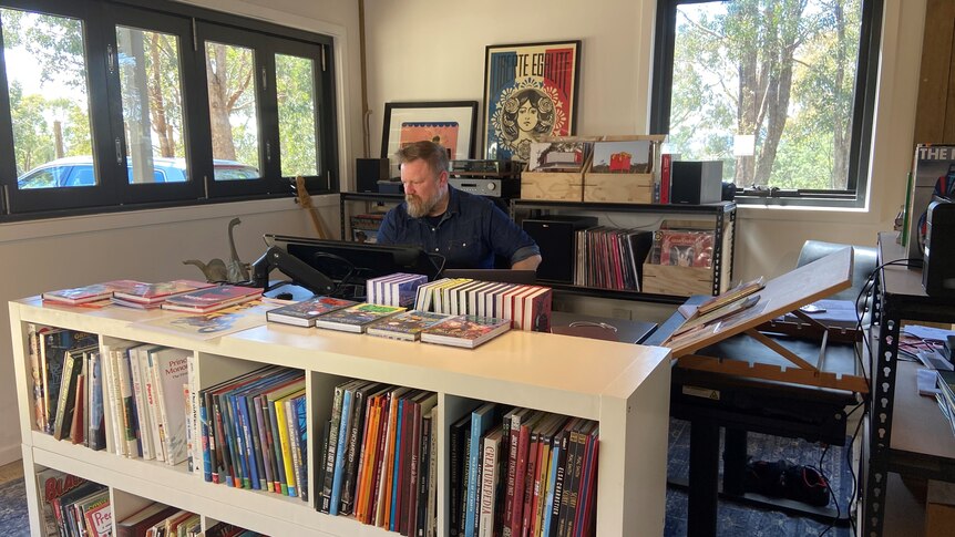 A man sitting at a screen behind a white bookshelf, with records and art in the background. He's in an outside studio.