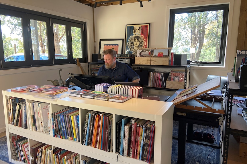 A man sitting at a screen behind a white bookshelf, with records and art in the background. He's in an outside studio.