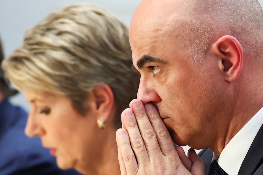 A man with palms pressed together in front of his face during press conference with woman out of focus beside him