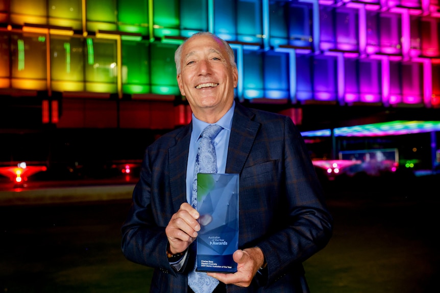A man with grey hair holds a glass award in front of a building lit up rainbow.