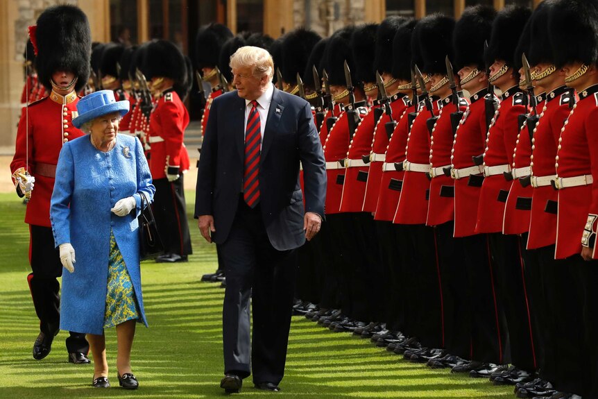 The Queen in a light blue coat and hat walks alongside Mr Trump, on the grass, passing a line of men in beefeater uniforms