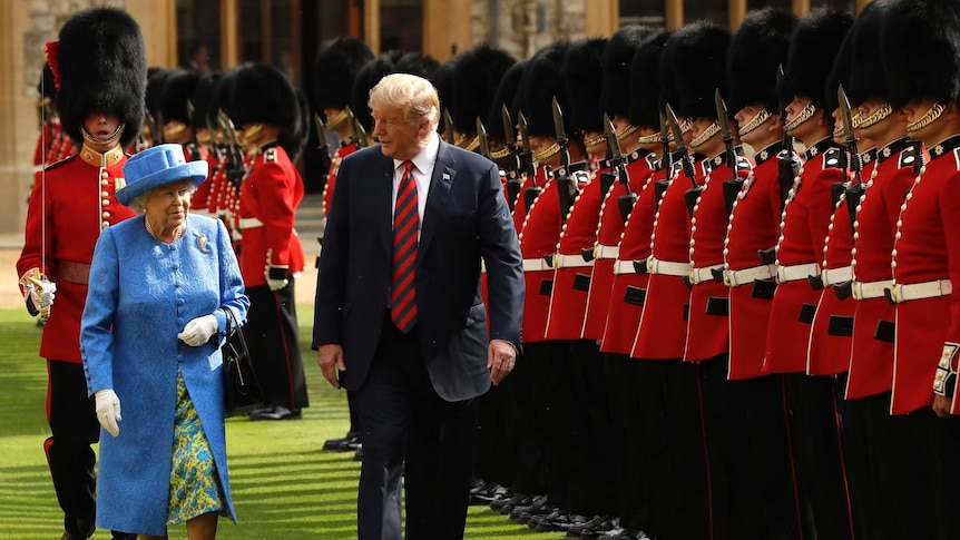 The Queen in a light blue coat and hat walks alongside Mr Trump, on the grass, passing a line of men in beefeater uniforms