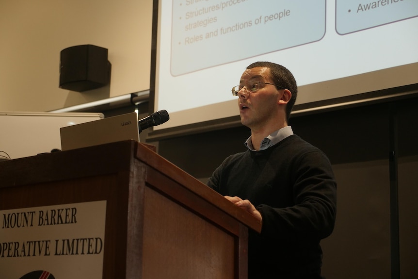 a man wearing glasses and a jumper speaks at a lectern