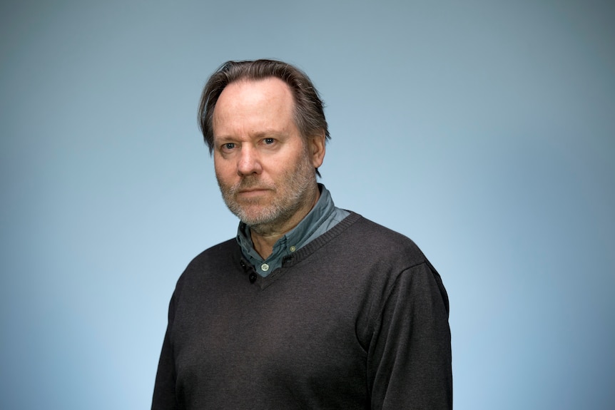 A man with a receding hairline and long hair sits before a blue screen