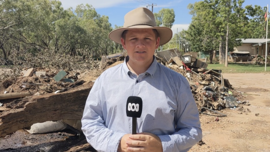 Man wearing a hat, holding ABC microphone with piles of rubbish behind him.