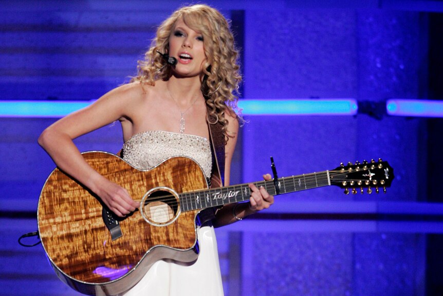 Taylor Swift plays on her guitar on stage while singing into a microphone.