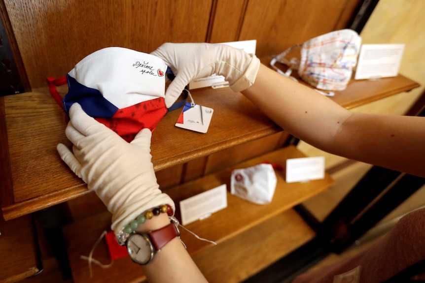 You view a pair of hands holding a face mask in the colours of the Czech national flag sitting on a brown wooden shelf.