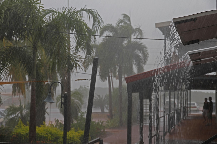 Rain drips off a building as two people stand undercover, looking out at palm trees, street lights and a shade sail