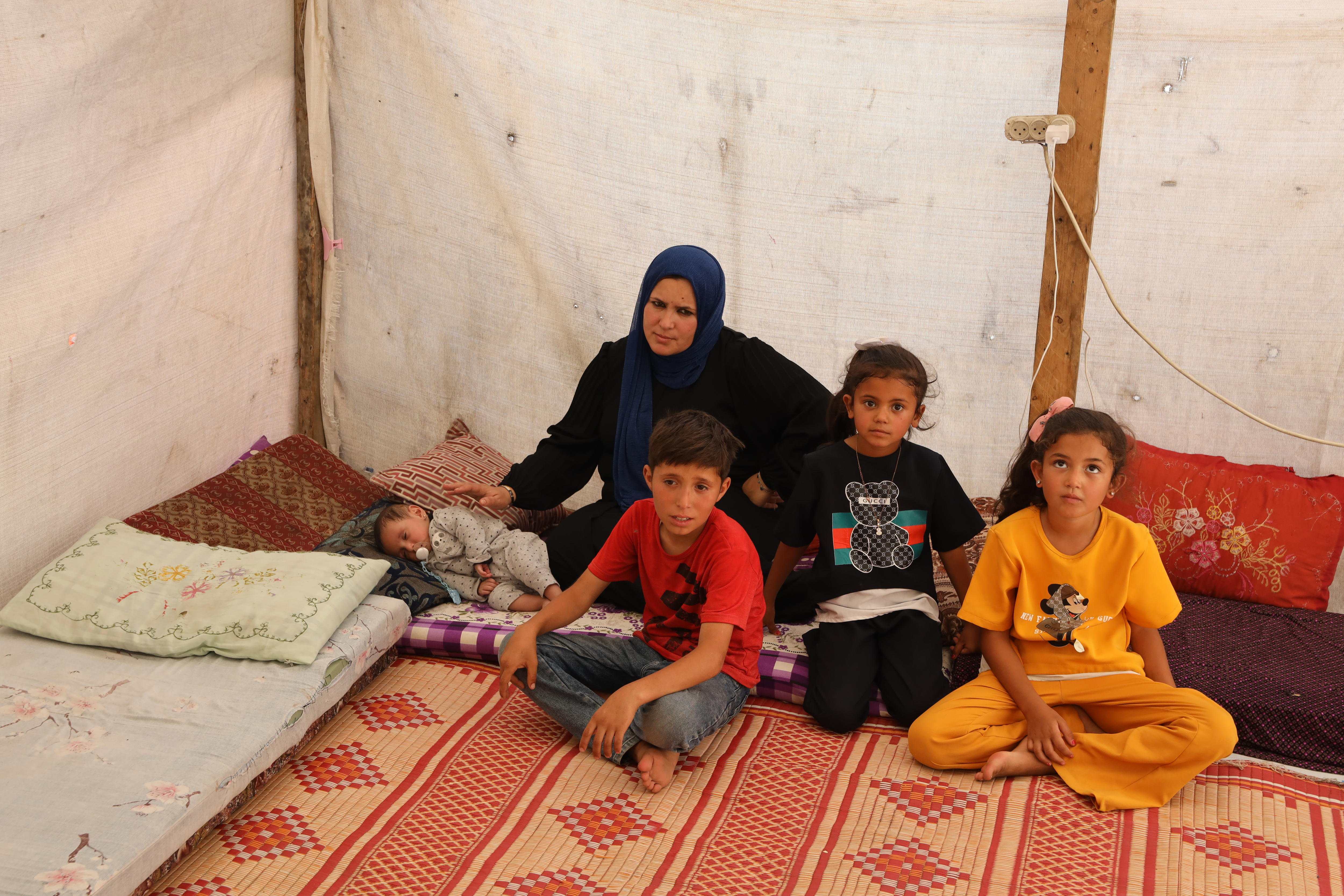 A mother wearing a hijab sits on a mat inside a tent with a baby and three children.